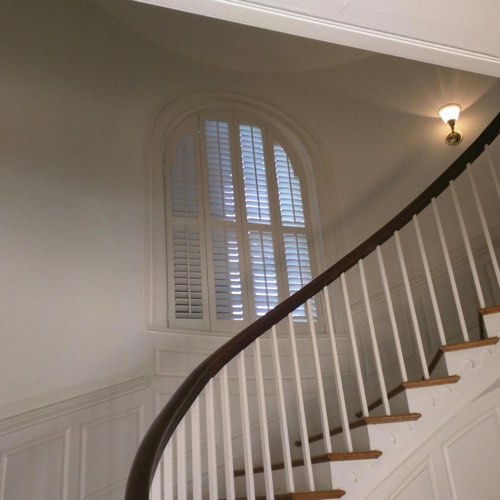 White plantation shutters covering rounded window located in curved stairwell.